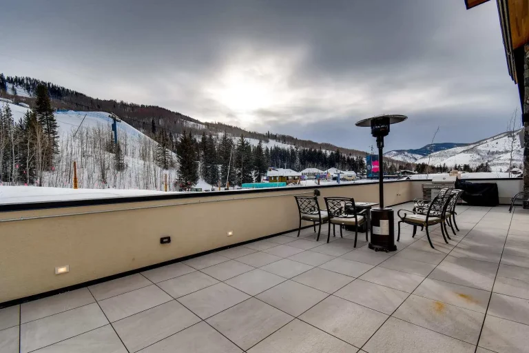 Outdoor terrace with patio furniture, a heater, and a BBQ grill overlooking snow-covered mountains and a ski resort.