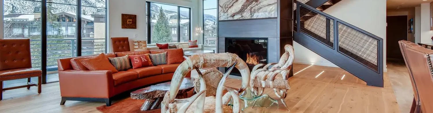 Modern living room with a curved orange sofa, wooden chairs, a glass coffee table, and a large fireplace under a staircase. Large windows offer a snowy mountain view.