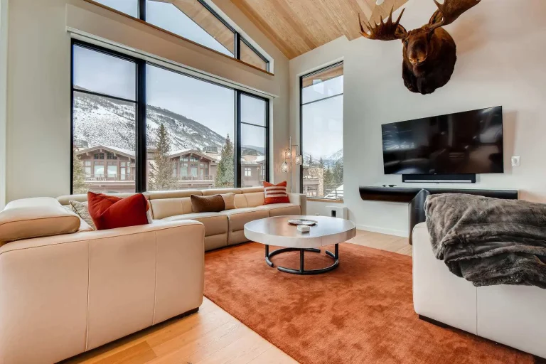 Modern living room with cream sofas, a large TV, a mounted moose head, floor-to-ceiling windows with a mountain view, and a round coffee table on an orange rug.