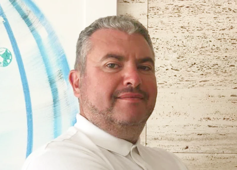 A man with short gray hair and a beard, wearing a white polo shirt, stands against a textured wall.