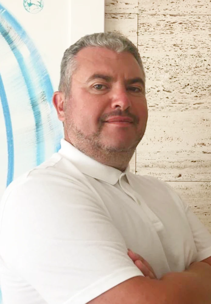 A man with short gray hair and a beard, wearing a white polo shirt, stands with arms crossed in front of a textured wall.