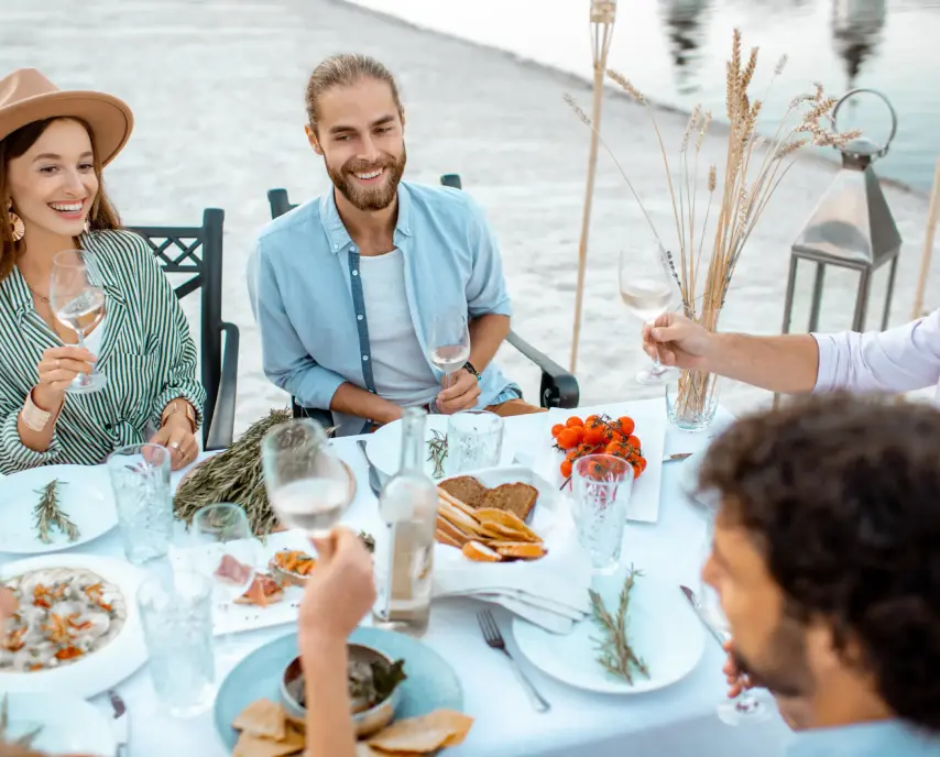 A group of people enjoy an outdoor meal, smiling and raising glasses, with a table set with various dishes and a scenic water view in the background.