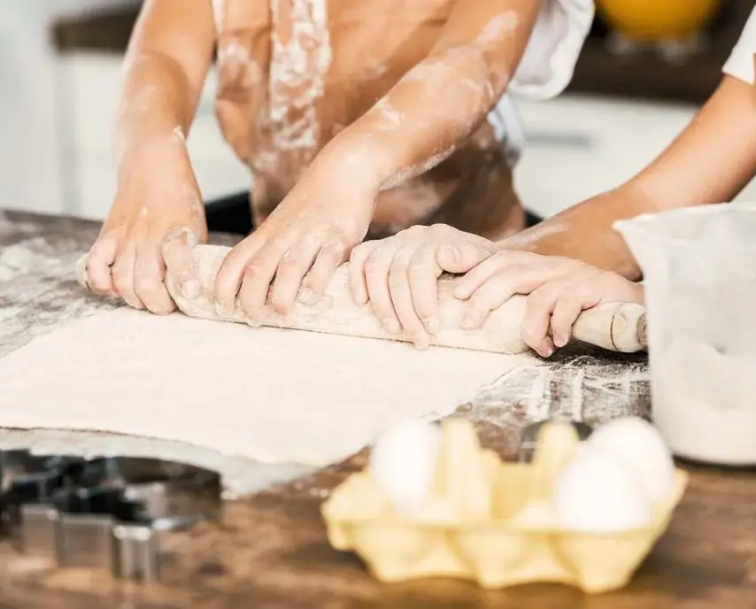Two people rolling out dough with a rolling pin on a floured surface, with eggs and flour in the foreground.