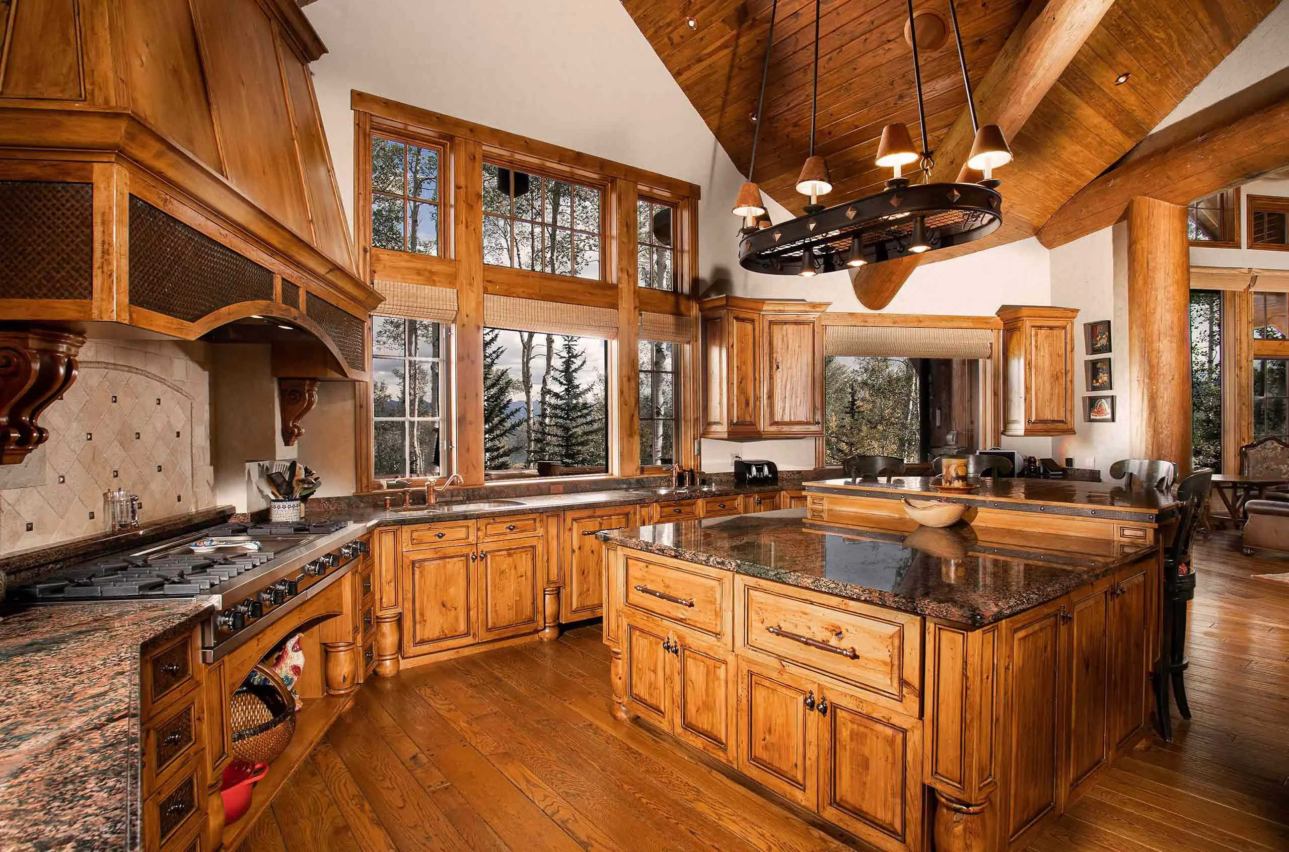 Rustic luxury kitchen with wooden cabinetry, granite countertops, and large windows overlooking a forested landscape.