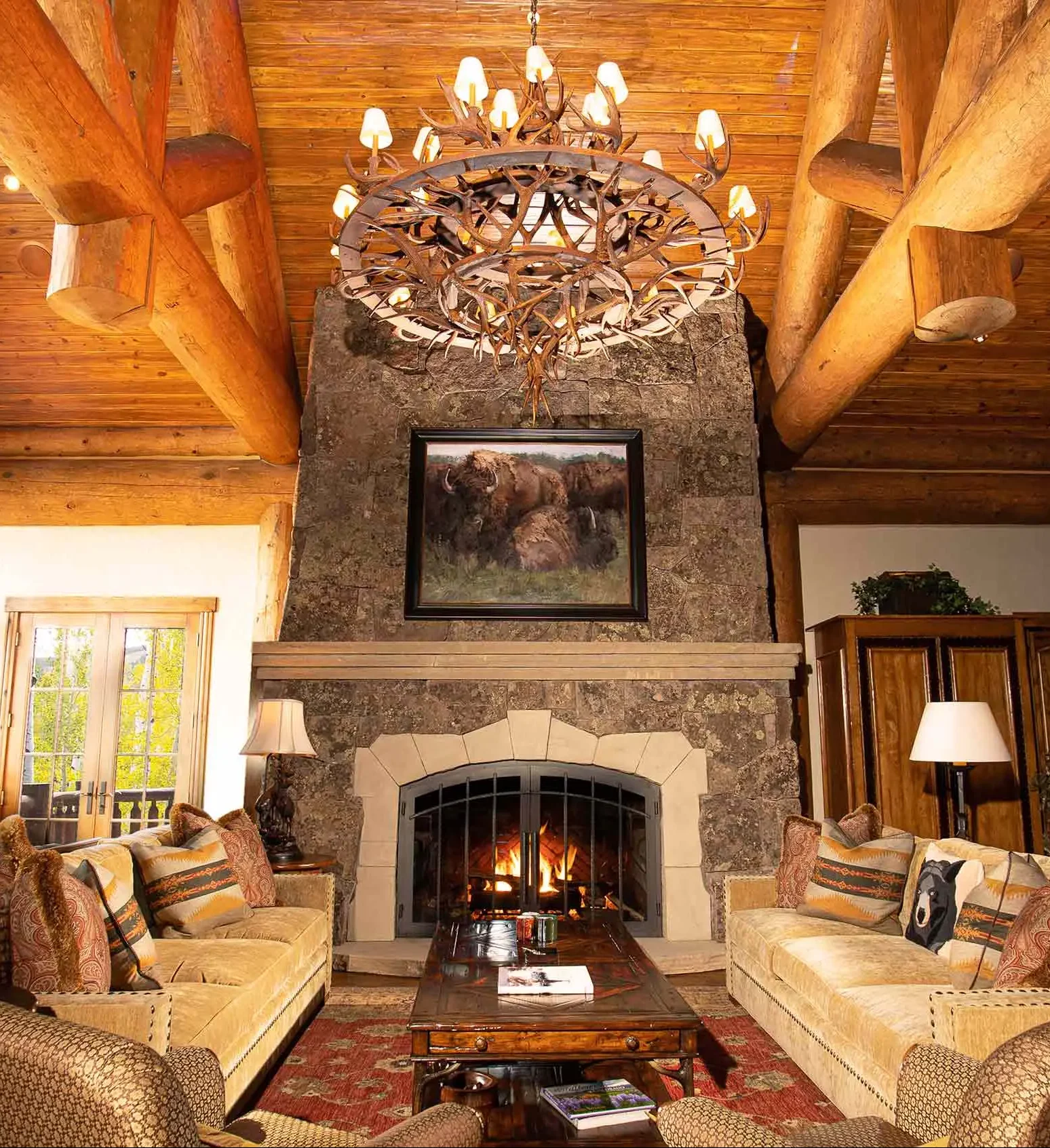 Rustic living room with a stone fireplace, beige sofas, a wooden coffee table, and a decorative chandelier.