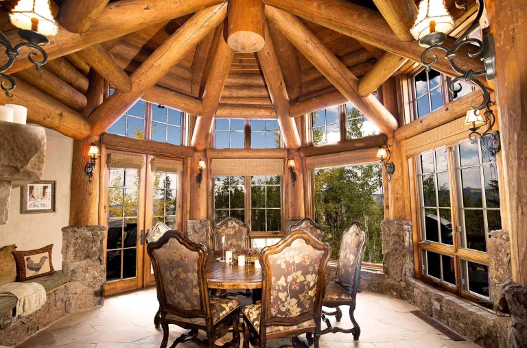 Rustic luxury villa interior with a round dining table and floral upholstered chairs, surrounded by large windows and wooden beams.