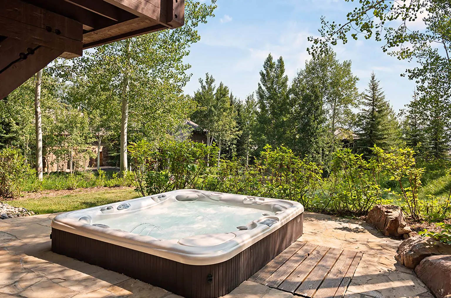 Outdoor hot tub on a stone patio surrounded by lush greenery and trees.