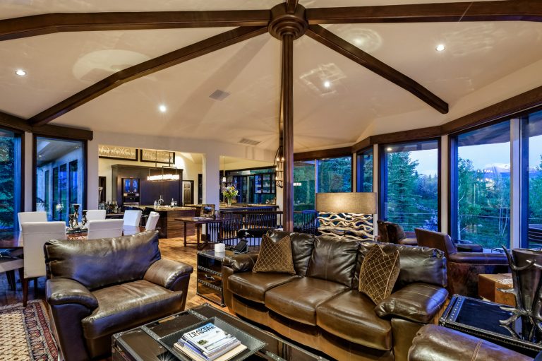 Luxury villa interior with leather sofas, wooden beams, large windows, and a view of the surrounding forest and mountains.