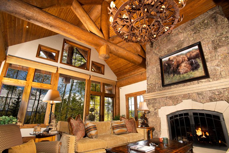 Rustic living room with wooden beams, large windows, a stone fireplace, and a cozy sofa with decorative pillows.