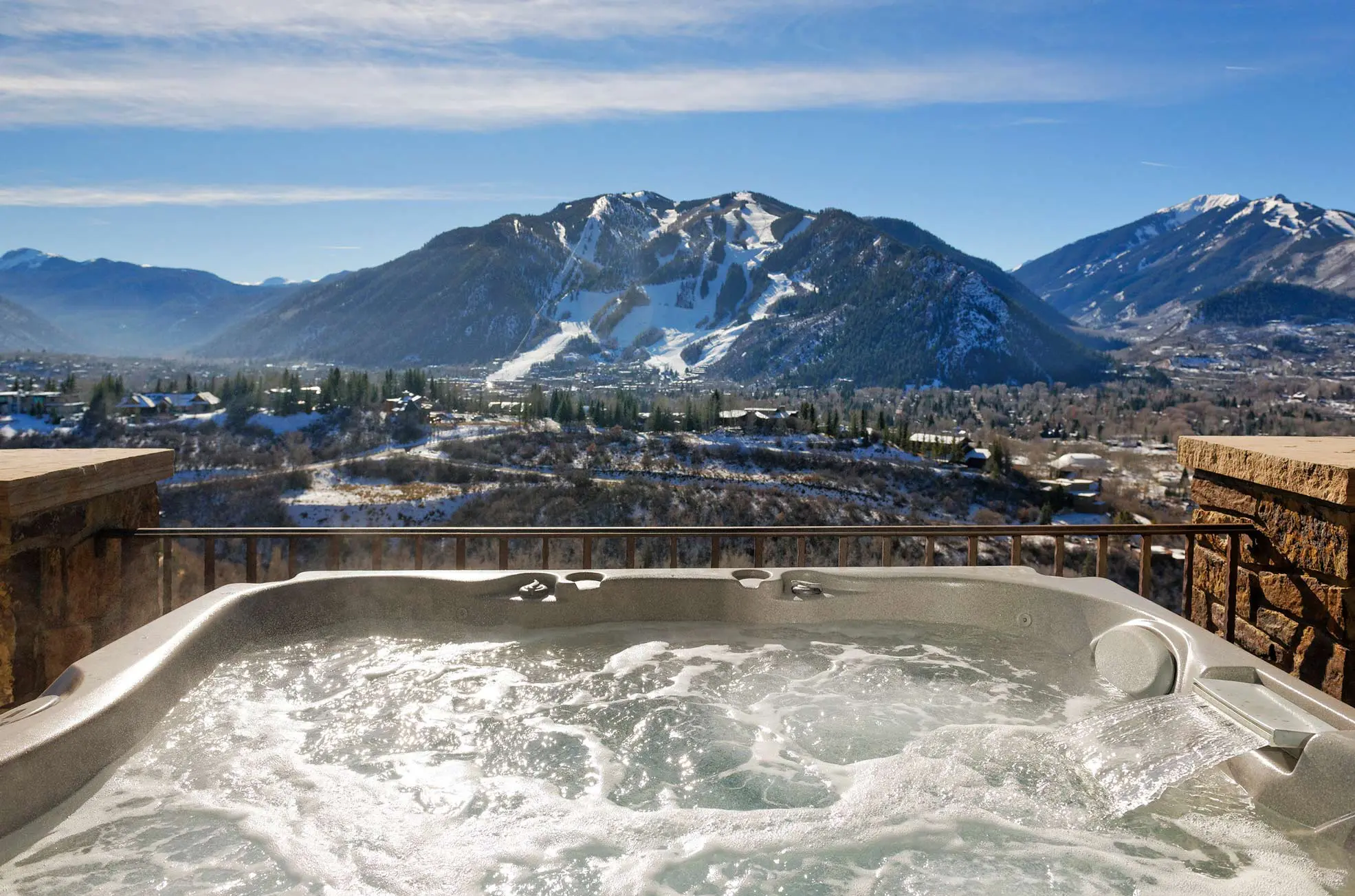 Outdoor hot tub with a view of snow-covered mountains and a ski resort in the distance.