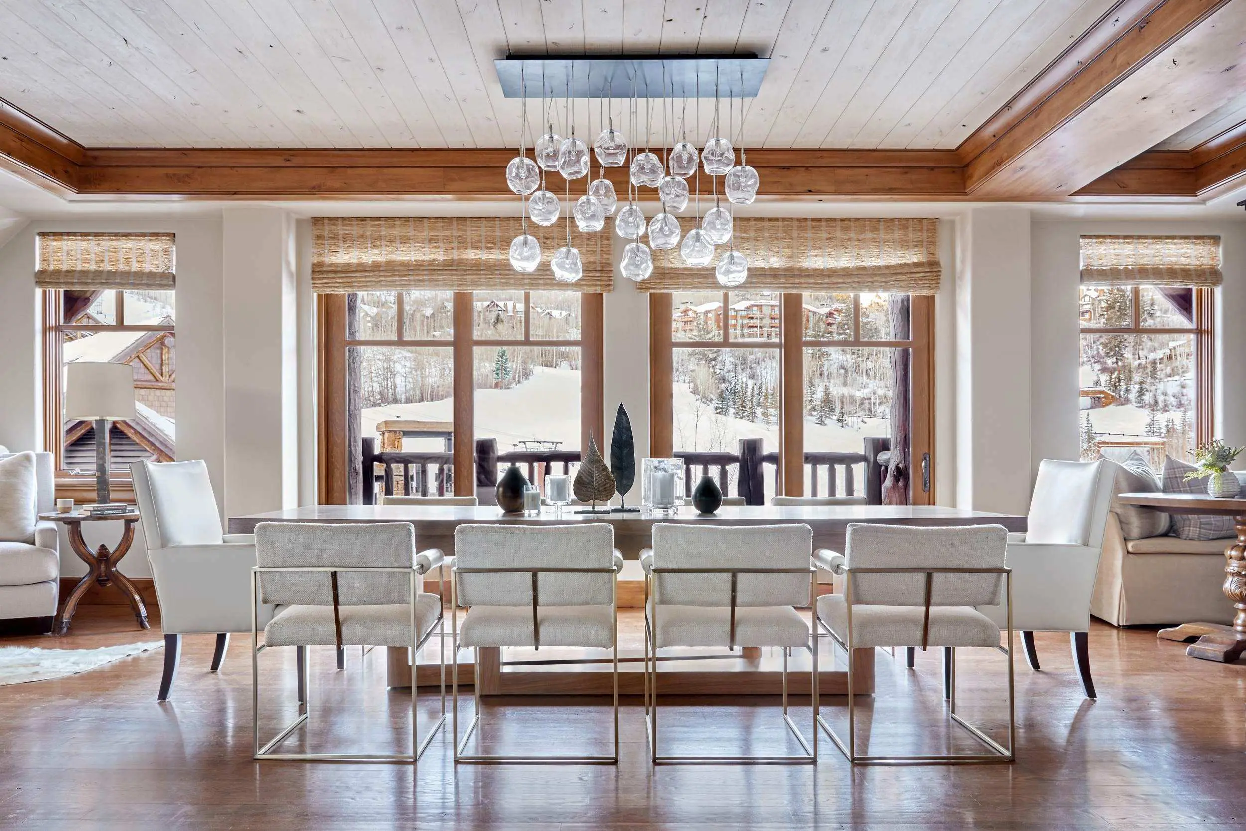 Luxurious dining room with a modern chandelier, large windows offering a snowy mountain view, elegant white chairs, and a wooden table.