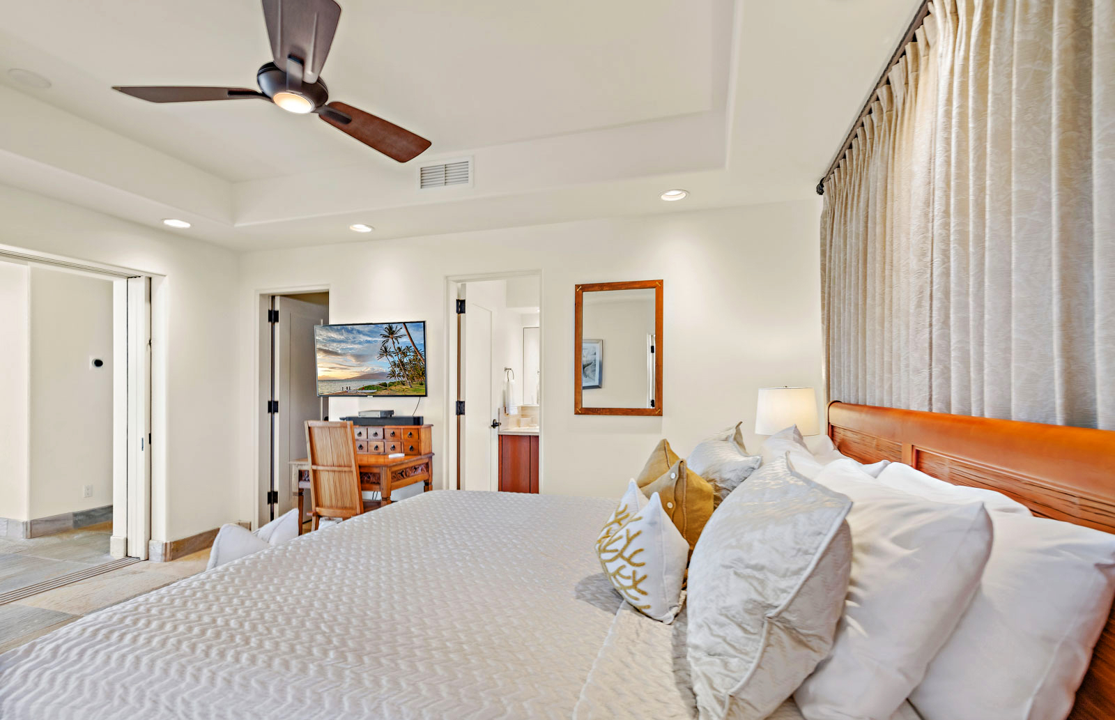 Modern bedroom with a large bed adorned with decorative pillows, a ceiling fan, a TV on a wooden dresser, and an open doorway leading to a bathroom.