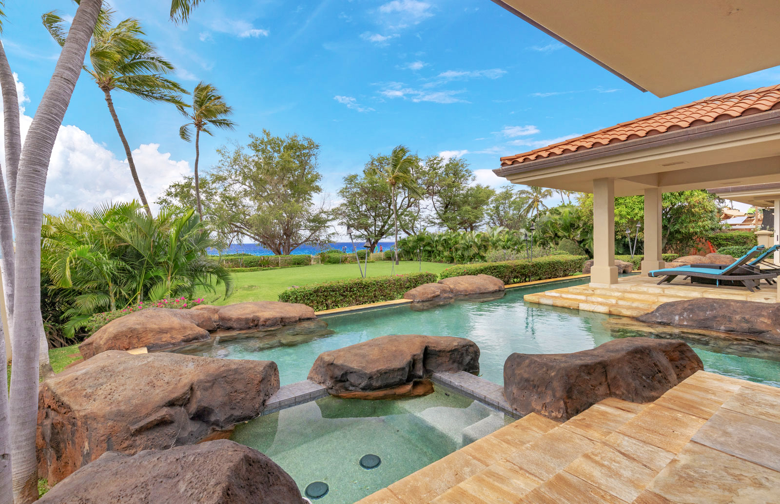 Luxurious outdoor pool area with stone accents, surrounded by lush greenery and palm trees, under a clear blue sky.