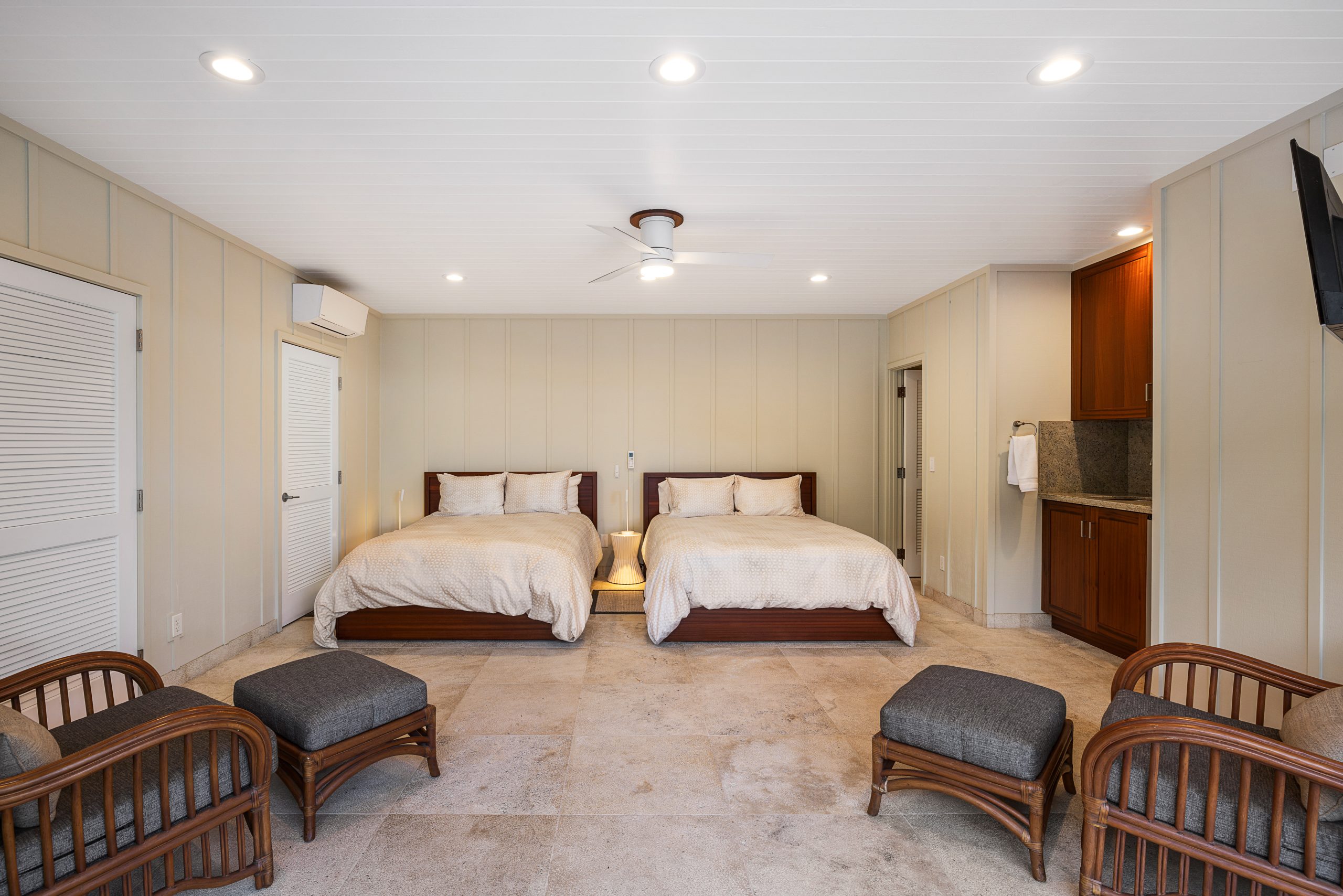 Hotel room with two double beds, a ceiling fan, wicker chairs with footrests, and a small kitchenette in the corner.