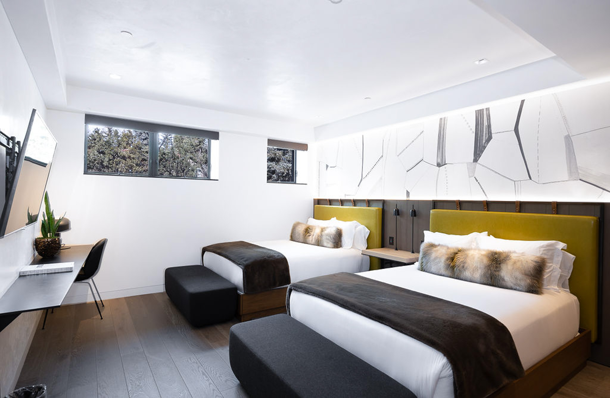 Modern bedroom with two large beds, contemporary artwork, desk, and small windows with a view of trees.