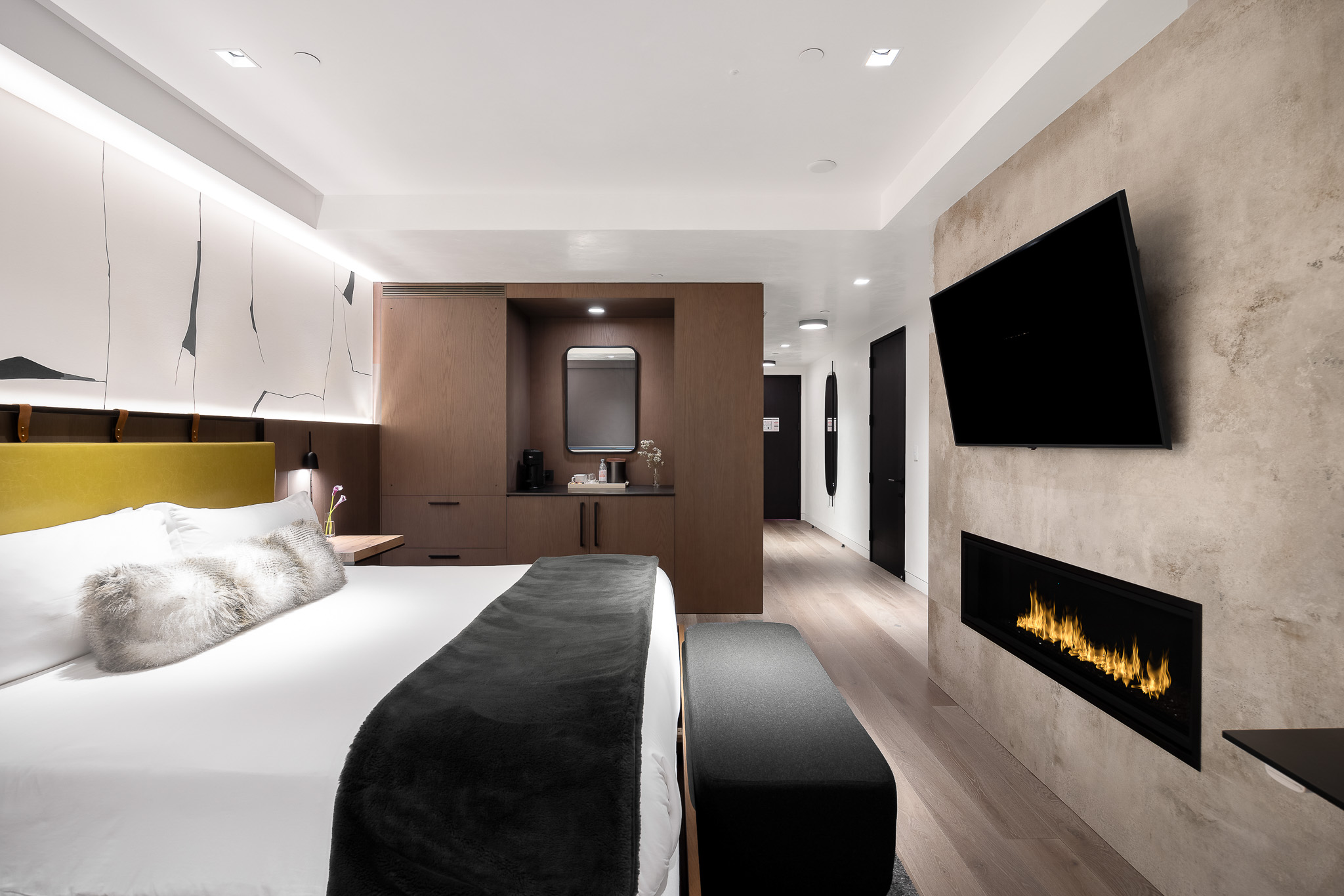 Modern luxury bedroom with a large bed, fur pillows, built-in fireplace, and wall-mounted TV. The room features minimalist decor with wood accents and soft lighting.