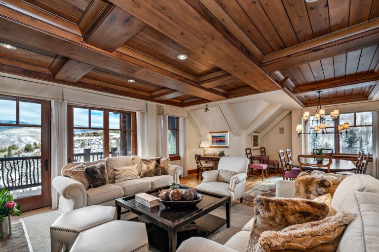 Luxury living room with wooden ceiling and large windows overlooking snowy mountains, featuring plush sofas, a coffee table, and a dining area with a chandelier.