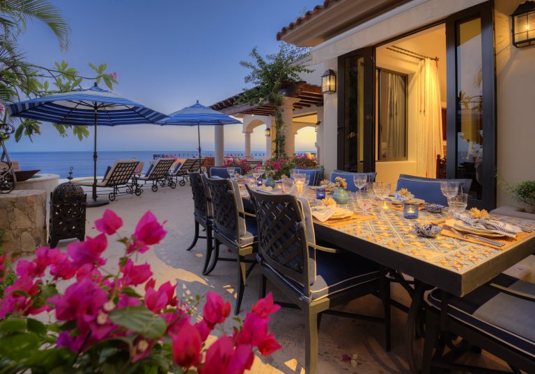 Outdoor dining area with a colorful table setting, surrounded by chairs and flowers, overlooking the ocean with lounge chairs and umbrellas.
