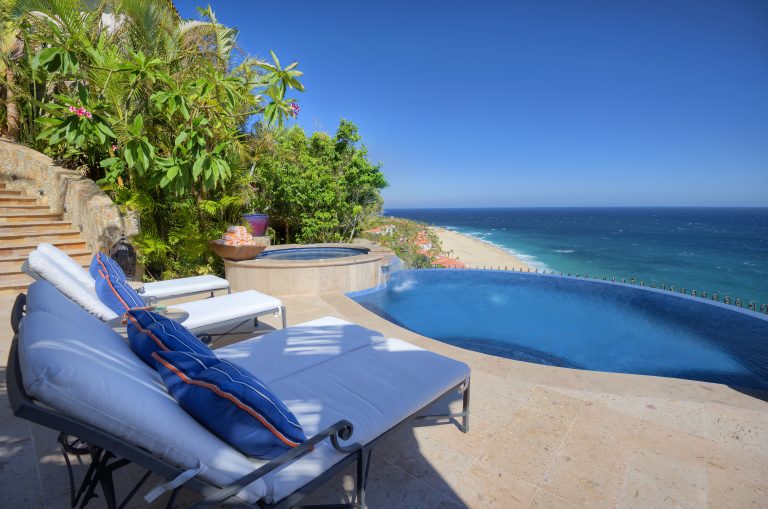 Luxury villa patio with loungers beside an infinity pool overlooking a beach and ocean, surrounded by lush greenery.
