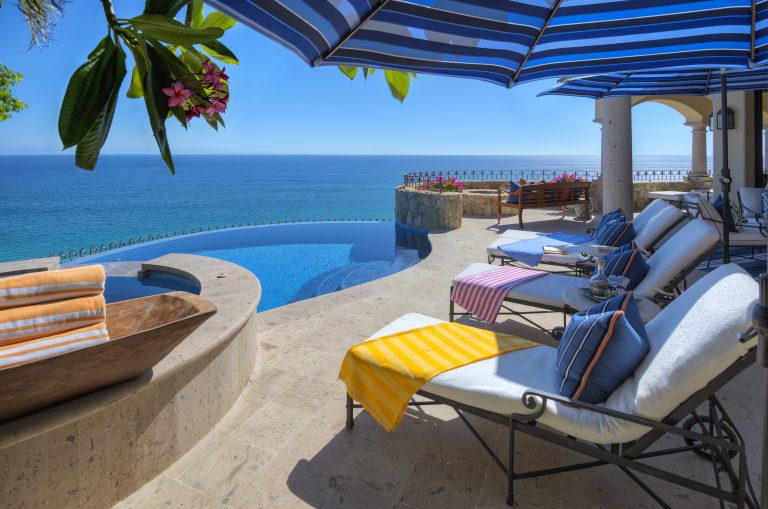 Outdoor patio with lounge chairs, striped towels, and umbrellas overlooking an infinity pool and the ocean.