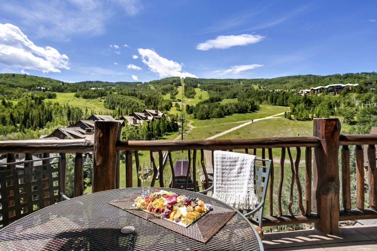 Scenic view from a balcony with a rustic railing, overlooking a mountain landscape and a table with a charcuterie board and wine glass.