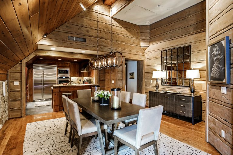 Rustic dining room with wooden walls, a large wooden table, modern lighting fixtures, and a view of a kitchen with stainless steel appliances.