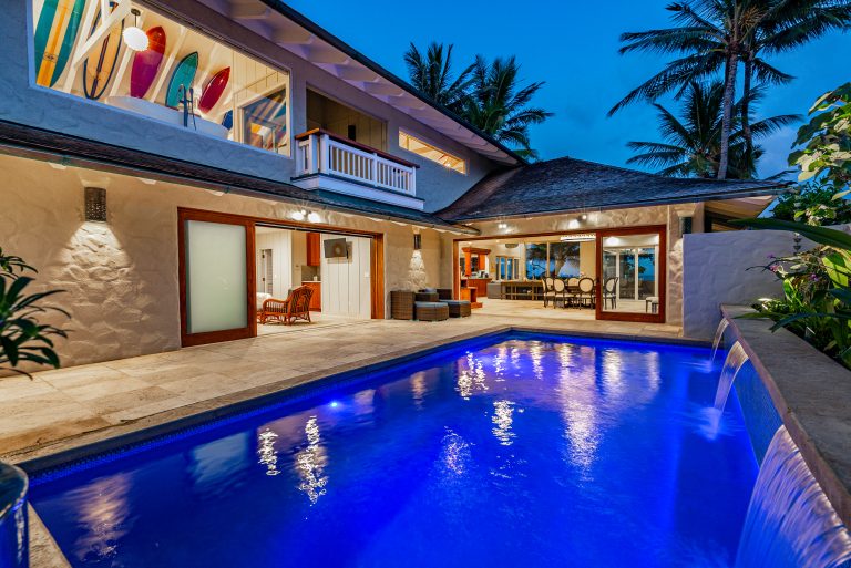 Luxury tropical house with illuminated swimming pool at dusk, featuring an open living area and surfboards displayed in an upper window.
