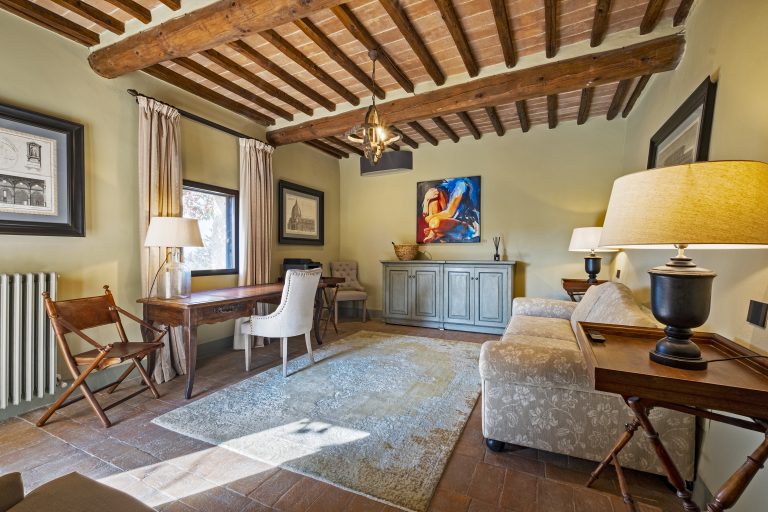 Cozy room with exposed beams, wooden desk, upholstered chairs, patterned sofa, artwork, and soft lighting.
