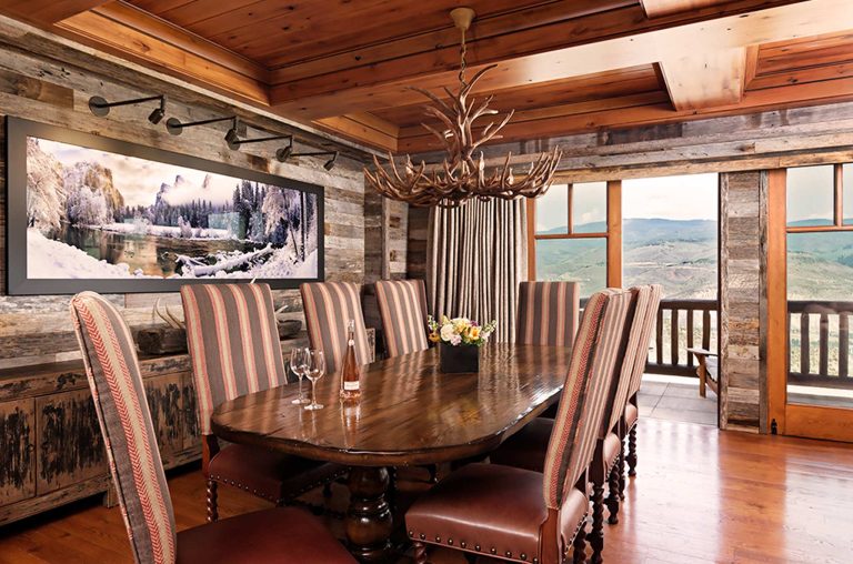Dining room with a rustic wooden table and striped chairs, antler chandelier, and large wall photo of a snowy landscape; mountain views from large windows.