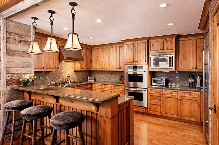 Rustic kitchen with wooden cabinetry, granite countertops, and hanging pendant lights, featuring a breakfast bar with three stools.