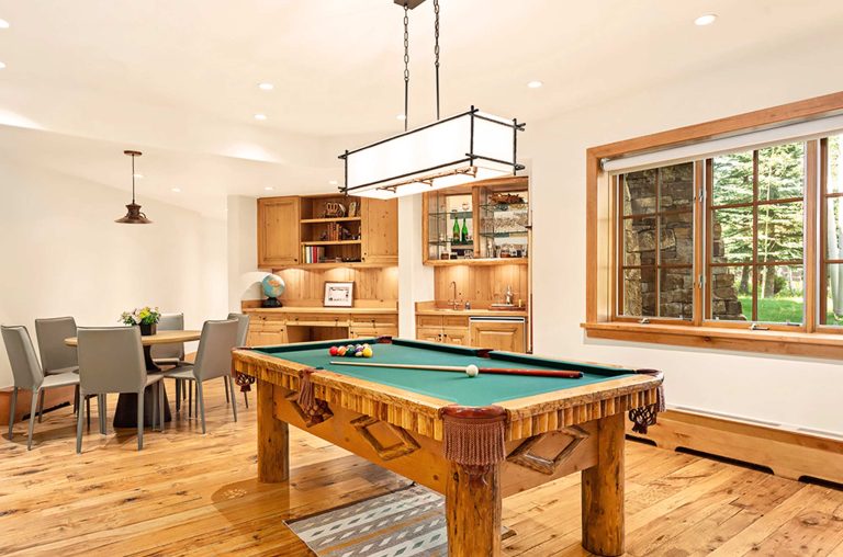A luxurious game room with a wooden pool table and a dining area with elegant chairs, featuring wooden flooring and cabinetry, and a large window with a view of trees outside.