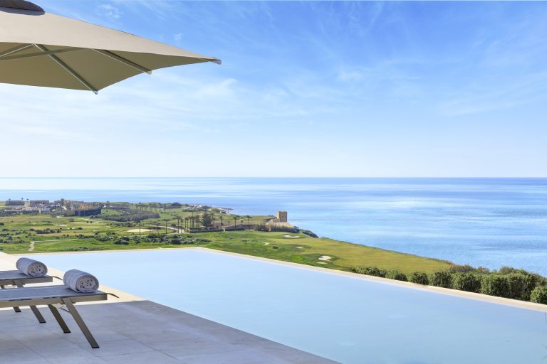 Infinity pool overlooking a coastal landscape with lush greenery, a golf course, and a tower near the ocean under a clear blue sky, partially shaded by an umbrella.