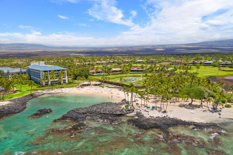 Aerial view of a luxury beachfront resort in Hawaii, surrounded by lush greenery and palm trees, with clear turquoise water and dark volcanic rock formations.