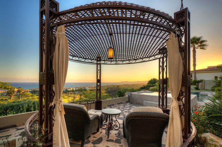 Outdoor seating area under a wrought iron gazebo with ocean view at sunset, overlooking a pool and lush landscape.