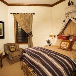 Cozy bedroom with rustic decor, including a patterned bedspread, armchair, drum-style side table, and a framed painting on the wall.