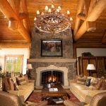 Rustic living room with a stone fireplace, log ceiling beams, large chandelier, and cozy furniture.