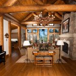 Rustic cabin-style living room with wooden beams, large stone fireplace, chandelier, and panoramic windows overlooking trees. Cozy seating area and upright piano in the foreground.