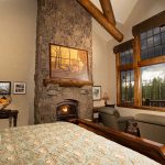 Cozy bedroom with stone fireplace, large windows overlooking the forest, patterned bedding, and comfortable seating.