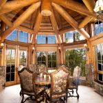 Rustic dining room with large log beams, wooden round table, decorative chairs, stone fireplace, and panoramic windows overlooking a forest.