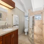 Modern bathroom with beige stone surfaces, a wooden cabinet, glass block shower, and various towels neatly hung.