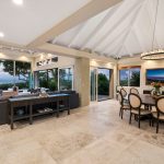Open-concept living and dining area with large windows offering ocean views, featuring a gray sectional sofa, round dining table with chairs, and a chandelier.