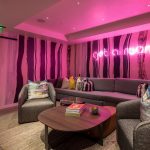 Luxurious lounge area with modern furniture, colorful pillows, abstract striped wallpaper, and a neon sign reading "get a room" in a luxury villa.