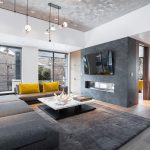 Modern living room with large gray sectional sofa, yellow cushions, marble coffee table, wall-mounted TV, and fireplace, featuring large windows with a view of snow-covered trees.