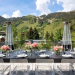Outdoor dining setup with floral arrangements, clear chairs, and patio heaters against a backdrop of lush green mountains in Aspen, Colorado.