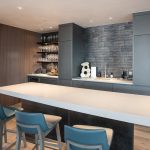 Modern kitchen bar area with sleek cabinets, a countertop with three chairs, open shelves displaying glassware, and a wall with textured tiles.