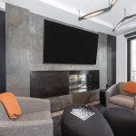 Modern living room with gray armchairs, orange accents, a fireplace, large TV, and a chess board on a black ottoman.