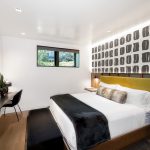 Modern bedroom with a king-sized bed, decorative fur pillows, abstract wall art, small desk with a chair, and large window.