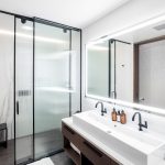 Modern bathroom with dual sink vanity, illuminated mirror, glass shower enclosure, and robe on a hook.
