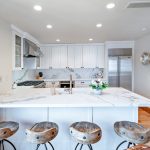 Modern kitchen with white cabinets, marble countertops, wooden bar stools, stainless steel appliances, and decorative wall pieces.