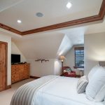 Bedroom interior with a large bed, wooden dresser, flat-screen TV, armchair, lamp, and a window with a mountain view.
